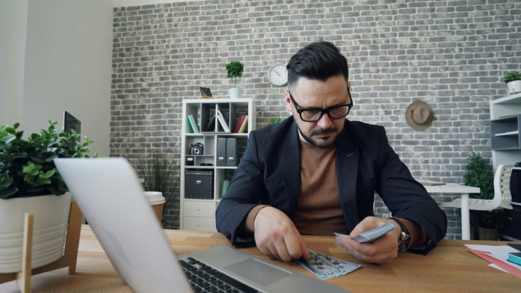 man counts cash in front of laptop in office setting