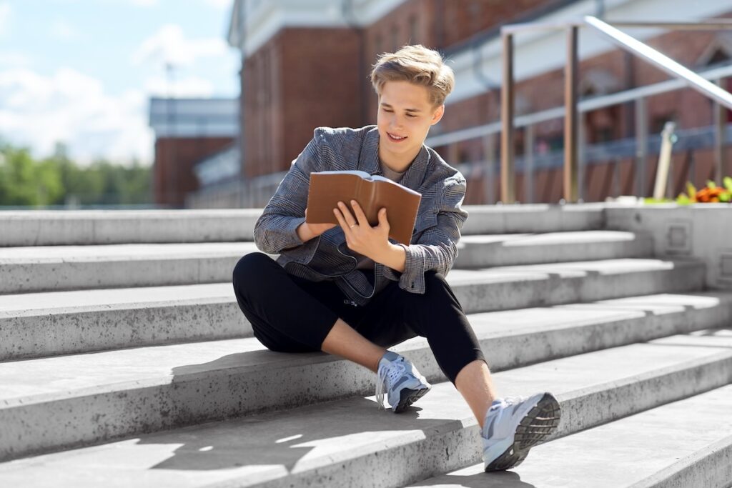 male university student sits on campus steps reading a book