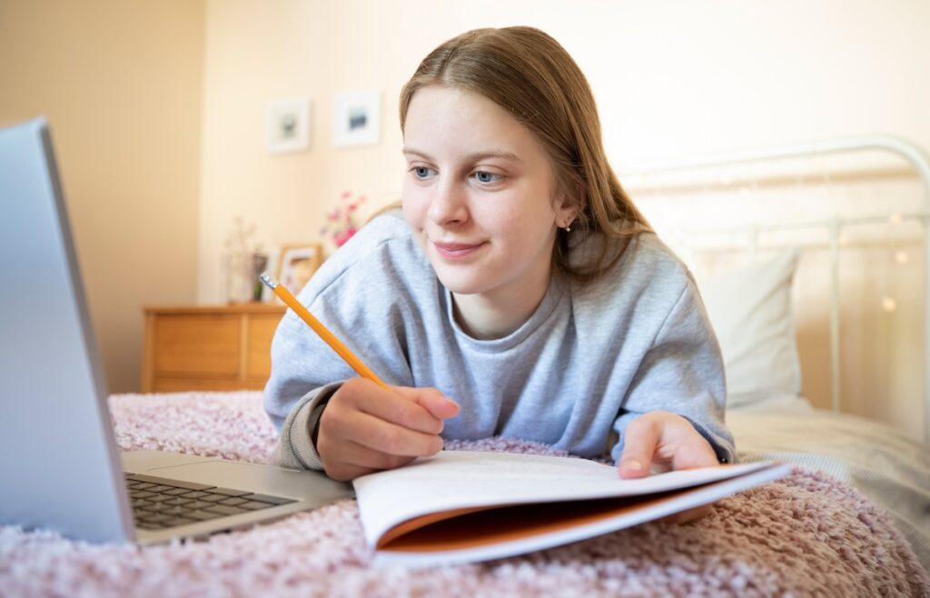 Teenage Girl Lying On Bed In Bedroom With Laptop Studying