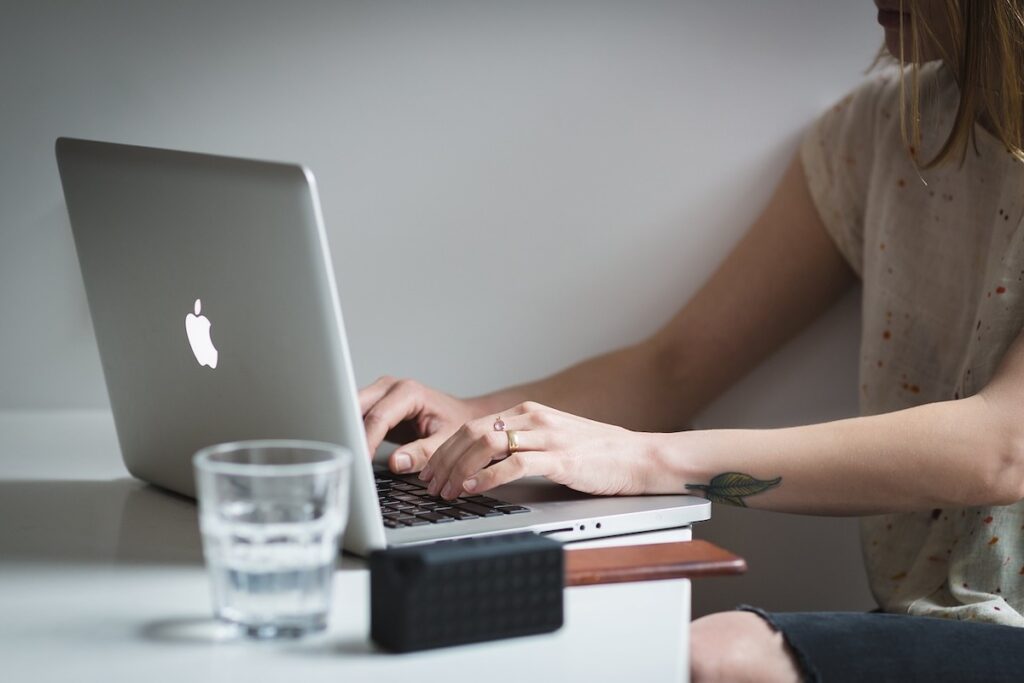 woman sits at desk typing on laptop with a glass of water next to her