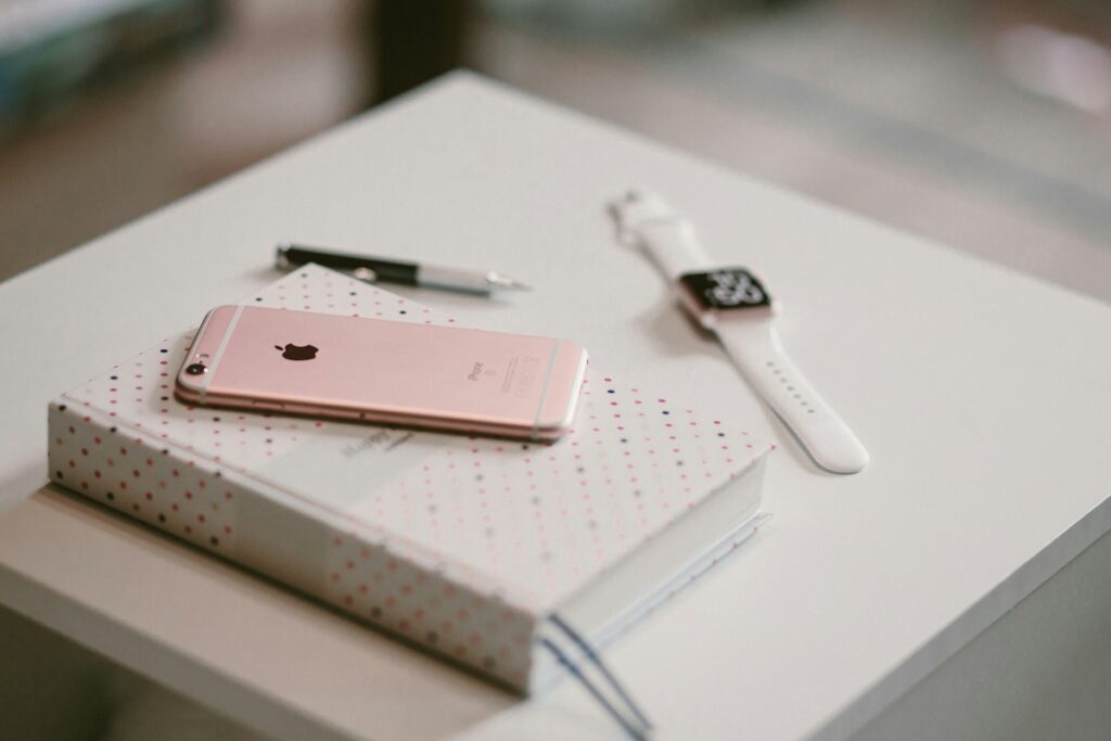 pink iphone sitting on top of notebook on white desk with an apple watch beside it