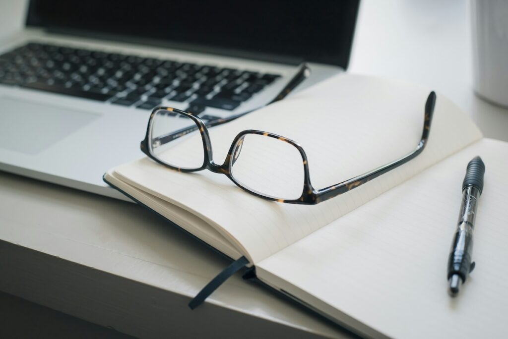 laptop with open notebook and pen next to it with glasses sitting on top