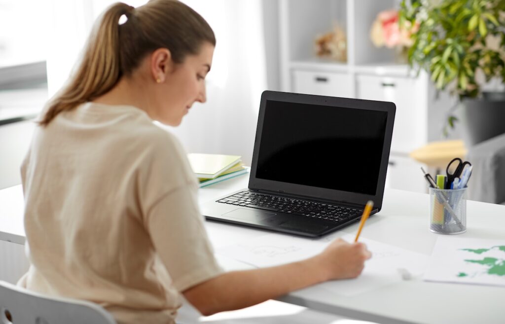 High school student drafting a college essay by hand beside a laptop, illustrating AI use in college essays.