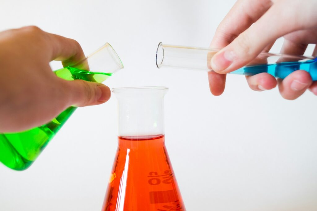 person pours green liquid from a test tube and blue liquid from another test tube into a jar holding red liquid