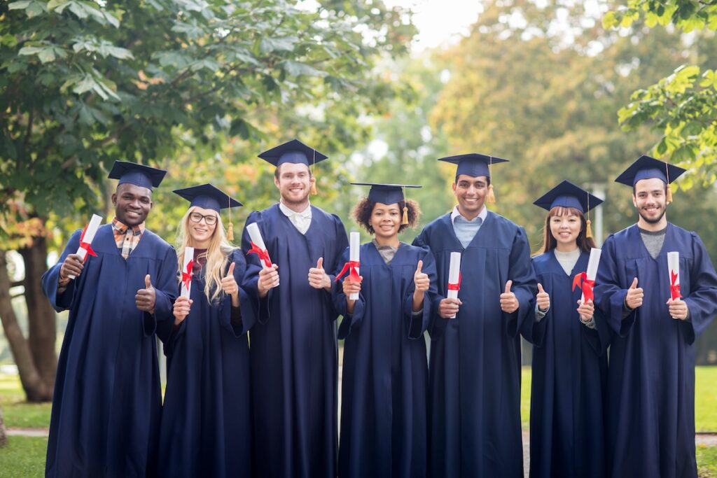 group of happy students in mortar boards and bachelor gowns with diplomas showing thumbs up