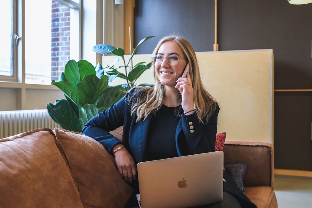 female student sits on leather sofa with a laptop on her lap while speaking on a cell phone