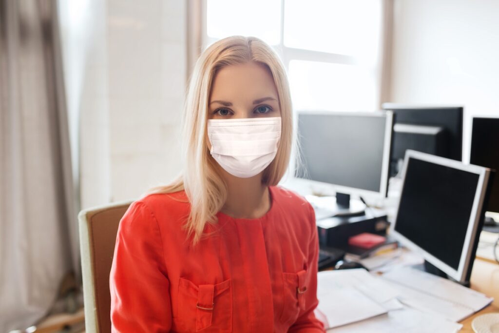 female office worker in mask sitting at a desk with multiple computers