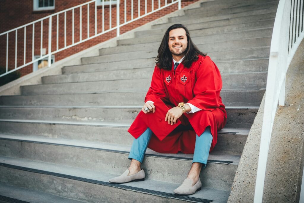 man in graduation cap and gown sits on steps at a college building