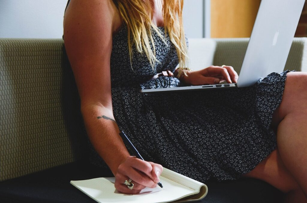 woman sits on couch with laptop in her lap and writing in notebook
