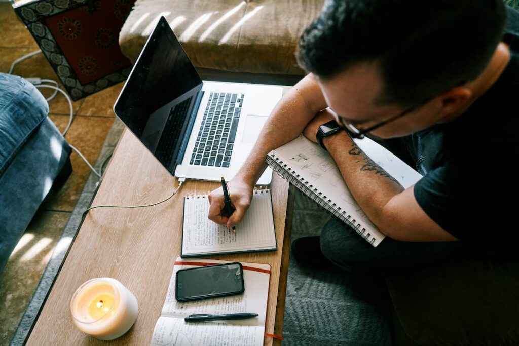 man works at laptop while writing notes on a notepad