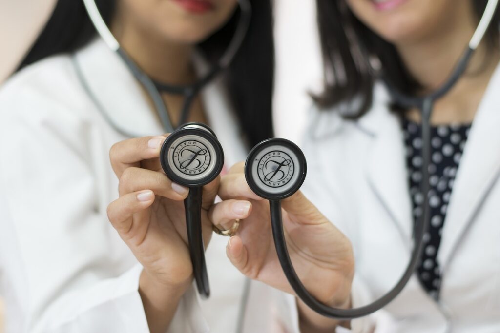 two women in medical lab coats hold stethoscopes up to the camera