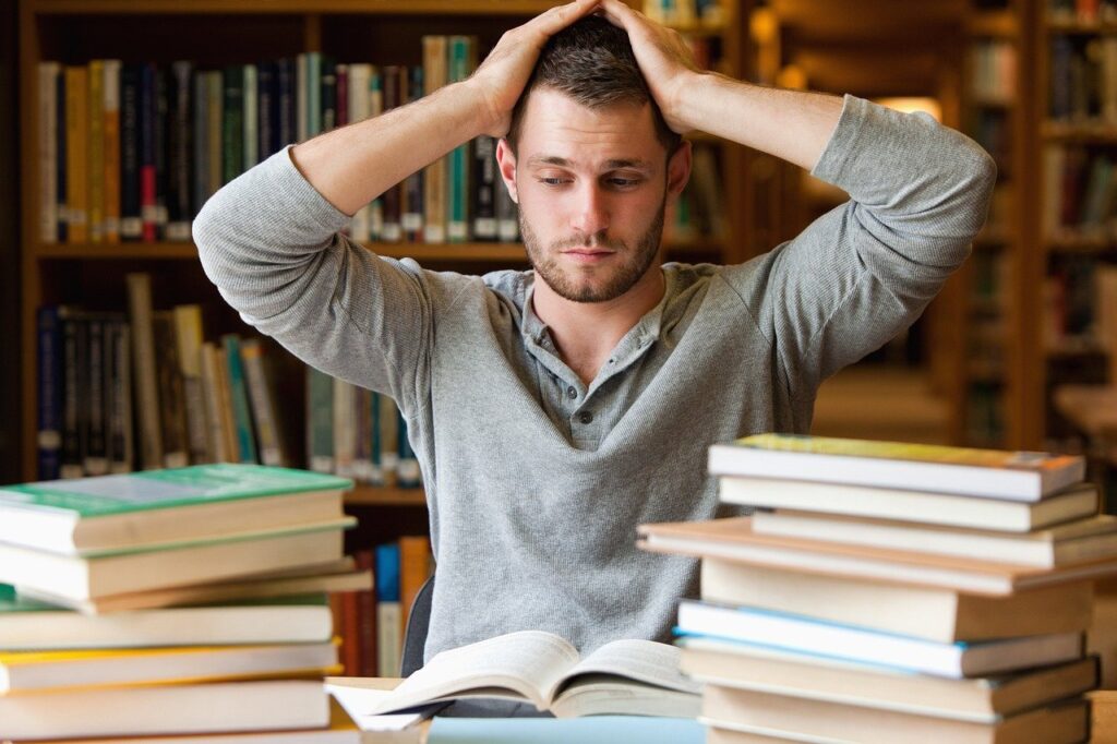 stressed student puts his hands on his head while looking at a pile of books in the library