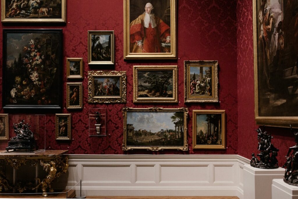 old paintings in frames hung on a red wallpapered wall in a museum