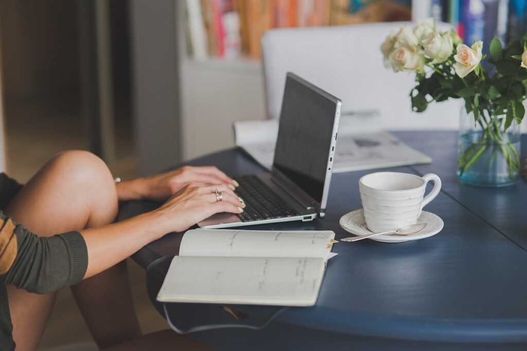 woman sits at table typing on laptop with an open notebook on the table next to her