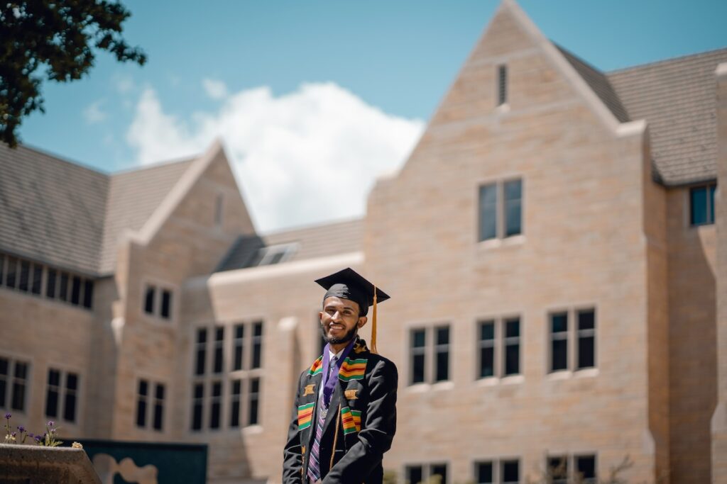 student in graduation cap and gown stands in front of university building