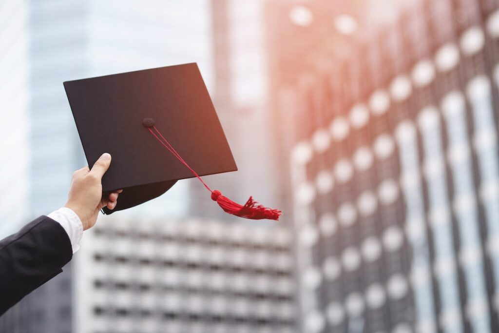 person holds graduation cap up with tall buildings in the background