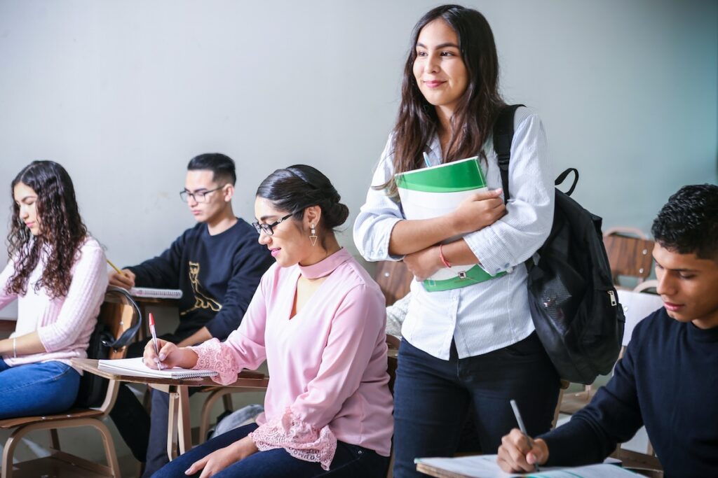 high school student stands with books in her arms while other students work at their desks