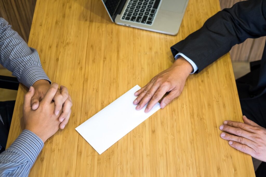 man slides envelope across the top of a wooden desk toward another man