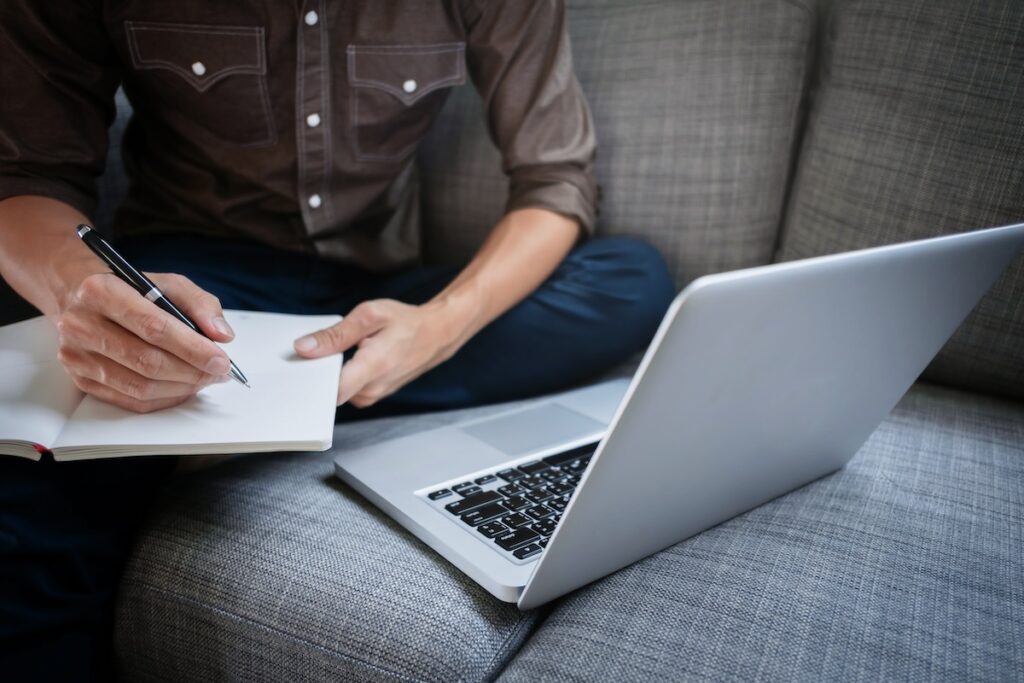 person working on laptop and writing on a notebook while sitting sofa