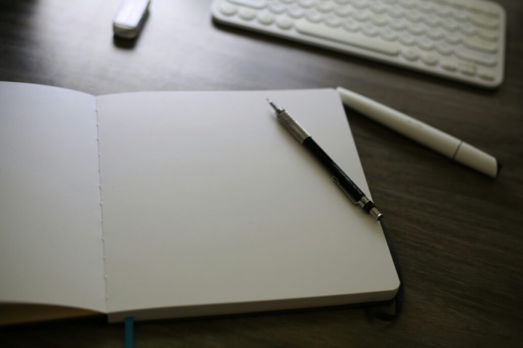 blank notebook on a desktop next to two pens and a computer keyboard