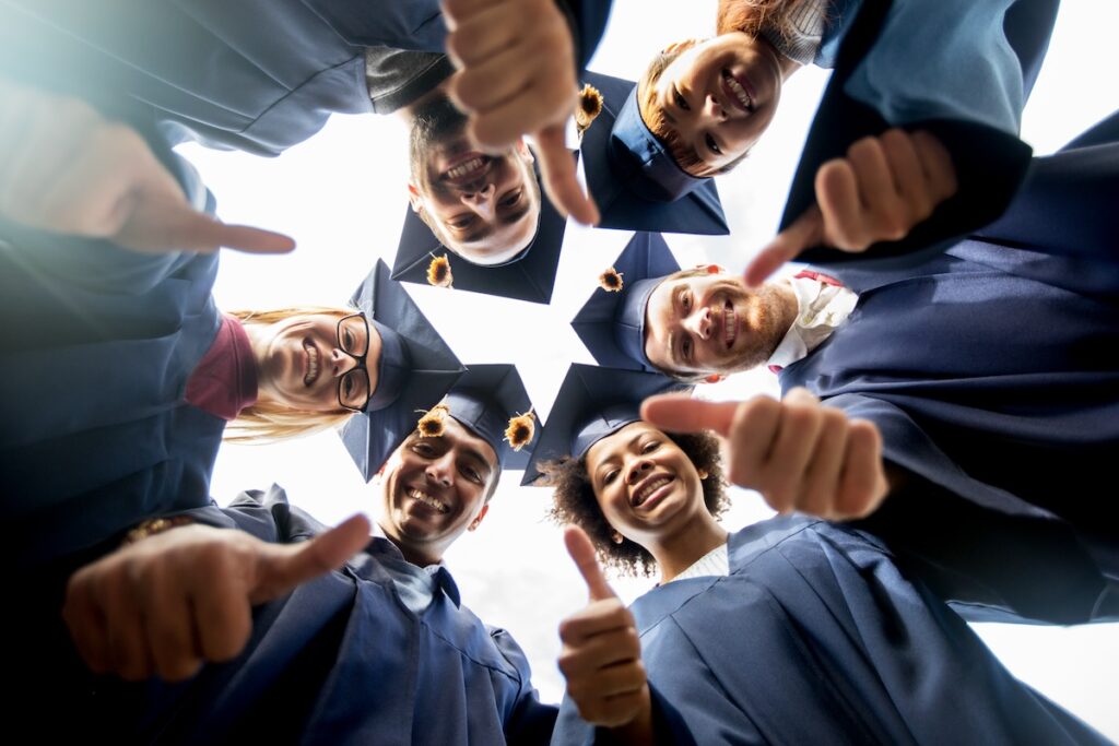 group of happy students in mortar boards and bachelor gowns standing in circle and showing thumbs up