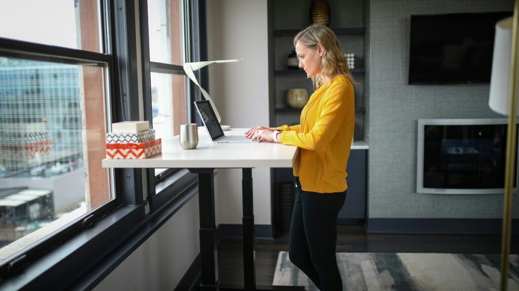 woman in yellow sweater working at laptop on standing desk in front of a window