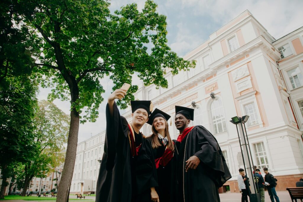 college students in graduation robes taking a selfie in front of university building