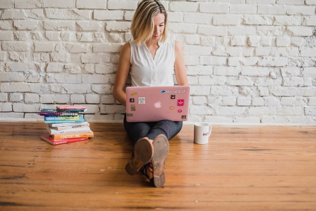 young woman sits on the floor working on a pink laptop in her lap with a stack of books next to her