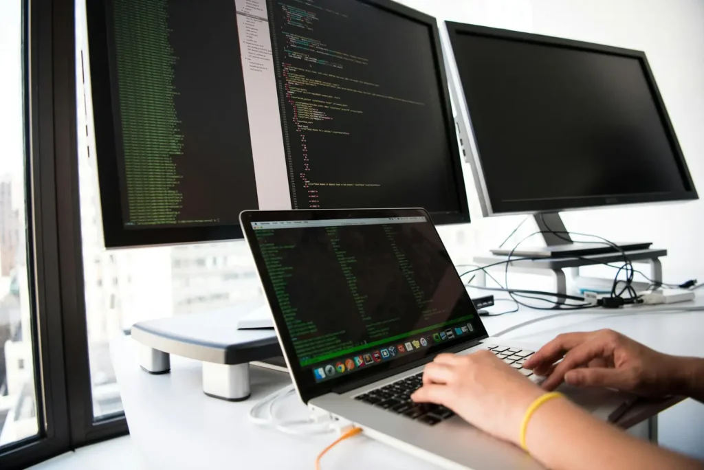 Best Colleges for Computer Science: A person's hands at a desk typing on a laptop with additional monitors showing code