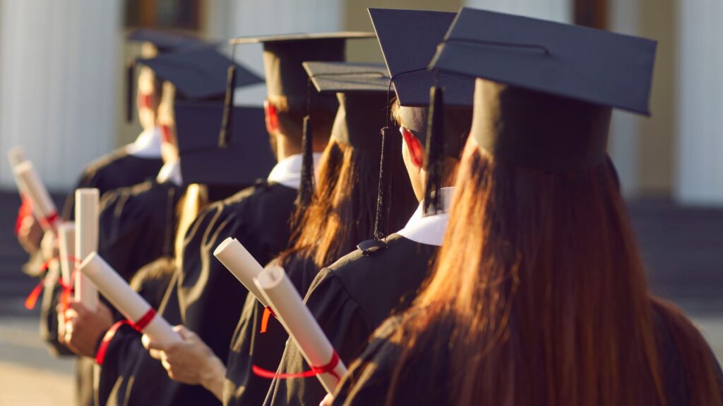High school students in full graduation regalia at a school graduation, viewed from behind, each holding a diploma with a red ribbon attached .