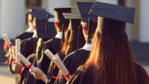 High school students in full graduation regalia at a school graduation, viewed from behind, each holding a diploma with a red ribbon attached .