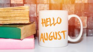 Cup of coffee with the words Hello August written in orange letters. Three books stacked on the left side of the image.