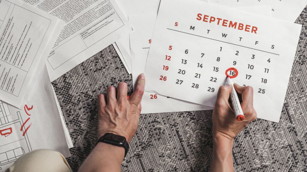Several printed papers on the floor being marked up by a person using red marker. Includes a September calendar.