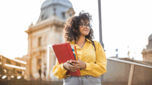 Woman wearing yellow blouse stands on a college campus with books in hand.