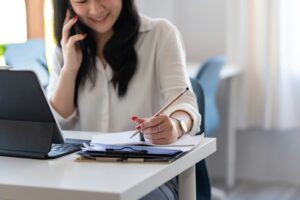 Woman smiling while talking on the phone taking notes on a piece of paper with a tablet screen open in front of her